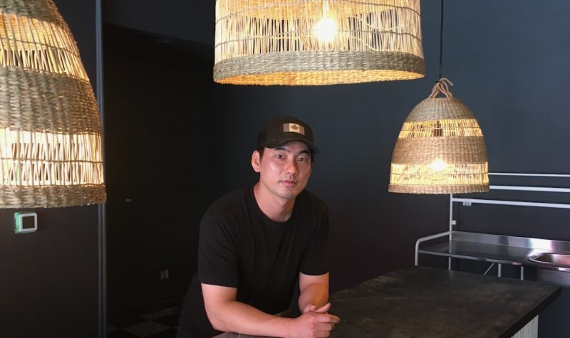 A young man in a ball cap leans on a black counter with rattan lighting hanging around him, and black walls behind him