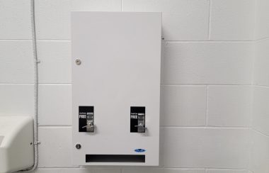 White metal dispenser with one tampon knob and one pad knob mounted on a white wall