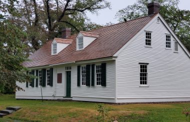 The outside of a white historic home on an overcast day with trees behind it and a yard in front.