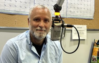 Man with greying hair and dark complexion in light blue shirt sits beside radio station microphone