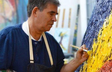 Peter W Hart portant tablier noir, chandail bleu devant une toile aux couleurs vives et pinceau à la main