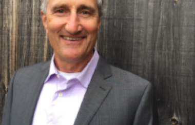 A man in a grey suit against a wooden background outside.