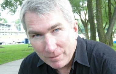 Man posing for picture wearing a black shirt. Trees and building sit in the background of the picture.