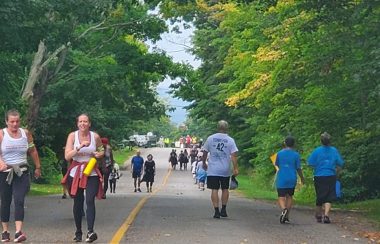 On peut voir quelques dizaines de marcheurs qui participent à la« Journée Terry Fox au cœur de la Baie Georgienne». Les participants sont de tous les âges et marchent sur une route entourée d'arbres.