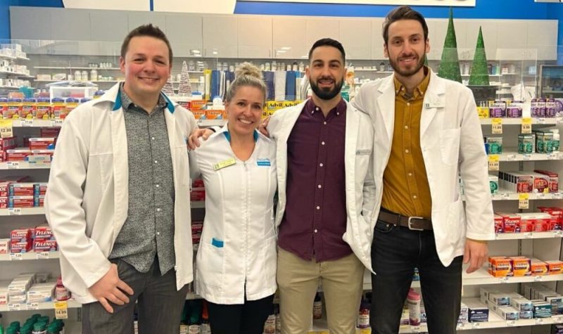 De gauche à droite, un pharmacien cheveux châtains, chemise grise, pantalon gris et sarrau blanc et bleu à l'effigie de Brunet. Pharmacienne blonde portant un sarrau blanc et bleu à l'effigie de Brunet. Pharmacien cheveux noir, barbe noir portant une chemise bourgogne, pantalons beiges et sarrau blanc et bleu à l'effigie de Brunet. Pharmacien cheveux châtains, barbe de même couleur que ses cheveux portant une chemise jaune, un pantalon noir et un sarrau blanc et bleu à l'effigie de Brunet. Ils sont devant un laboratoire inscrit le mot ordonnances en haut.