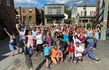 Pictured are members of the Sutton community after completely their flash mob downtown. It is a large group shot of participants with their hands in the air with some of Sutton's old buildings pictured in the background.