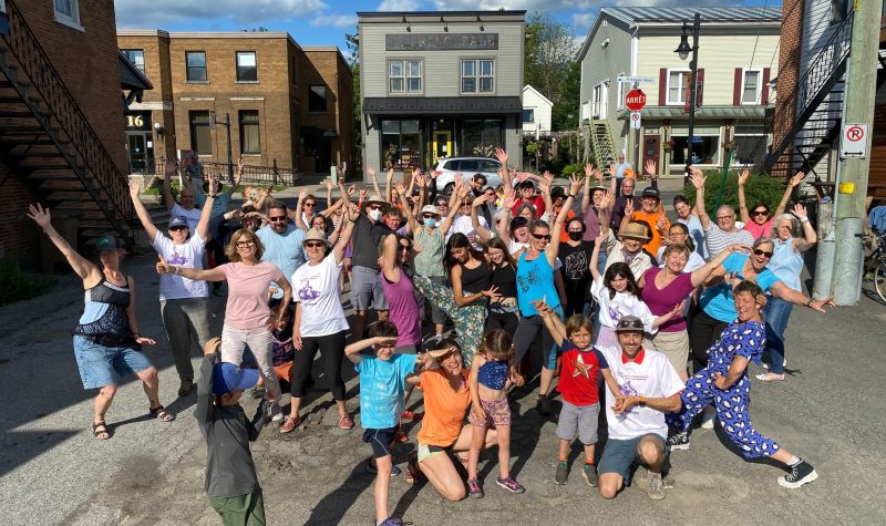 Pictured are members of the Sutton community after completely their flash mob downtown. It is a large group shot of participants with their hands in the air with some of Sutton's old buildings pictured in the background.