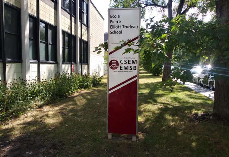L'enseigne de l'École Pierre-Elliott-Trudeau entre l'édifice et un arbre.