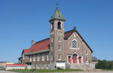 Grande église de couleur grise avec toiture et porte rouge. Fond de ciel bleu sans nuage.