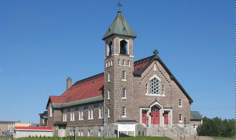 Grande église de couleur grise avec toiture et porte rouge. Fond de ciel bleu sans nuage.
