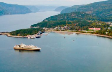 Paysage extérieur d'un ciel dégagé, de plusieurs montagnes au loin, le Fjord du Saguenay au centre. Il y a une bateau blanc et le village de Tadoussac vue de haut.