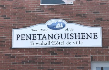 On voit l'affiche de l'hôtel de ville de Penetanguishene sur le mur de brique du bâtiment. Le symbole de la ville qui est un voilier blanc est visible et la mention « hôtel de ville » est inscrite dans les deux langues officielles