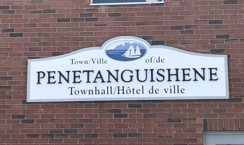 On voit l'affiche de l'hôtel de ville de Penetanguishene sur le mur de brique du bâtiment. Le symbole de la ville qui est un voilier blanc est visible et la mention « hôtel de ville » est inscrite dans les deux langues officielles