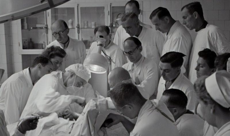 In a black and white photograph, a group of people, including doctors and nurses, stand in an operating room watching a doctor perform an operation. The clothes and hairstyles of the people in the room suggest the photo was taken in the mid-twentieth century.