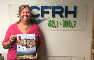 On peut voir Mme. Thérèse Fama souriante qui est photographier avec son livre intitulé « Who Knew, Growing up in Thunder Bay Beach » devant le logo de la radio de CRFH. La photo a été prise dans le studio de la radio.