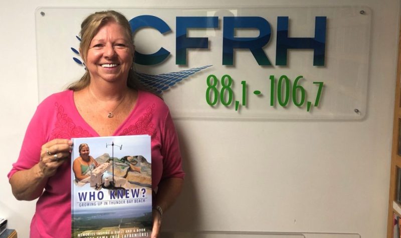 On peut voir Mme. Thérèse Fama souriante qui est photographier avec son livre intitulé « Who Knew, Growing up in Thunder Bay Beach » devant le logo de la radio de CRFH. La photo a été prise dans le studio de la radio.