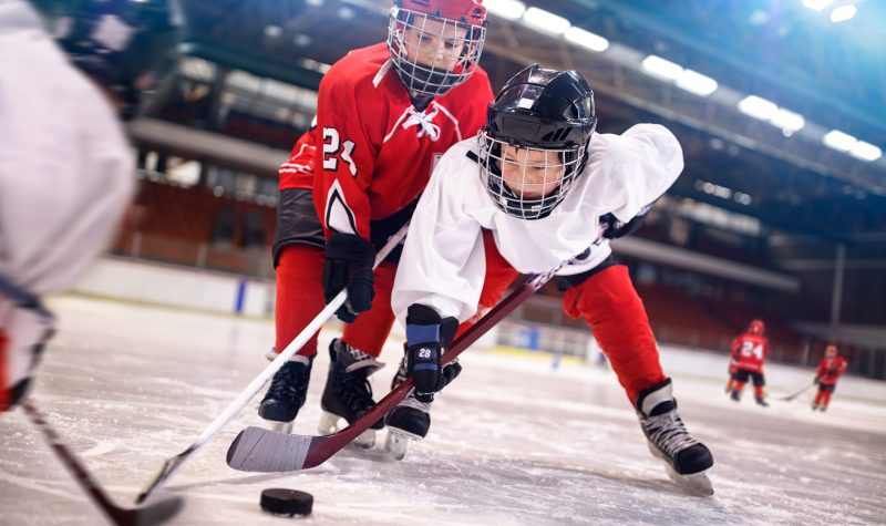 Deux jeunes joueurs de hockey, l’un à gauche avec un uniforme rouge et celui de droite uniforme blanc. Ils sont dans un aréna et les gradins sont derrière.
