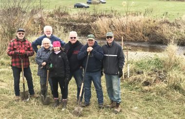 Seven of the volunteers that took part in tree planting along Quilliams stream. With shovels in hand, they stand in front of the stream.