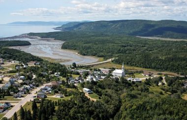 Vue d'ensemble de la municipalité des Bergeronnes. Montagnes au loin, fleuve, route 138, arbres, maisons et église.