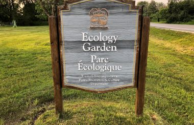 On voit le panneau indicatif du Parc écologique de Penetanguishene. Le panneau est en bois et l'identification est en anglais et en français. Le panneau est placé sur le gazon du parc près de la rue Fox.