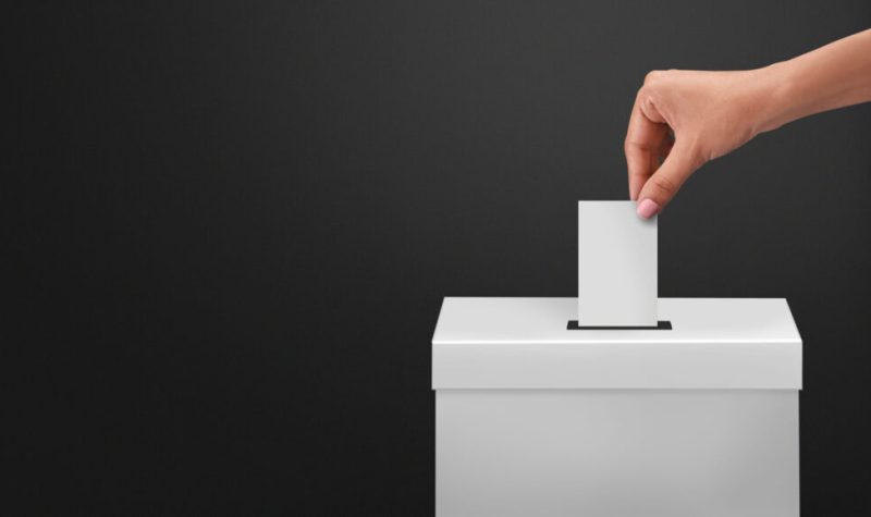 Main d'une femme allant porter son bulletin de vote blanc dans une boite blanche avec un mur gris foncé à l'arrière.