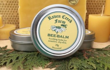 Photo promotionnelle d'un des produits à base de miel de Raven Creek Farm. Il s'agit d'un baume pour la peau dans un contenant rond avec une étiquette ronde sur le dessus du contenant, de couleur jaune avec l'image d'une abielle sur une marguerite et le logo de Raven Creek Farm.
