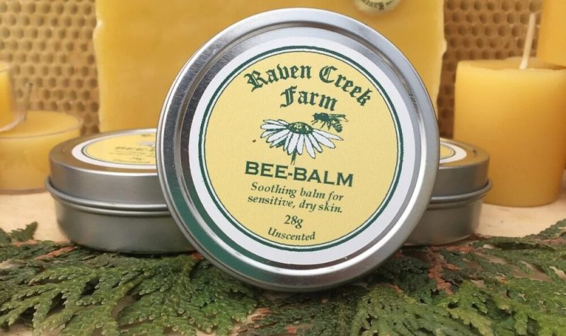 Photo promotionnelle d'un des produits à base de miel de Raven Creek Farm. Il s'agit d'un baume pour la peau dans un contenant rond avec une étiquette ronde sur le dessus du contenant, de couleur jaune avec l'image d'une abielle sur une marguerite et le logo de Raven Creek Farm.