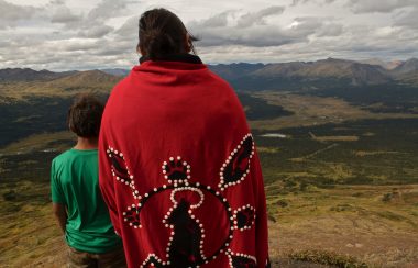 A woman in a red traditional indigenous blanket overlooking a valley with a young boy in a green shirt. both are facing away from the camera.