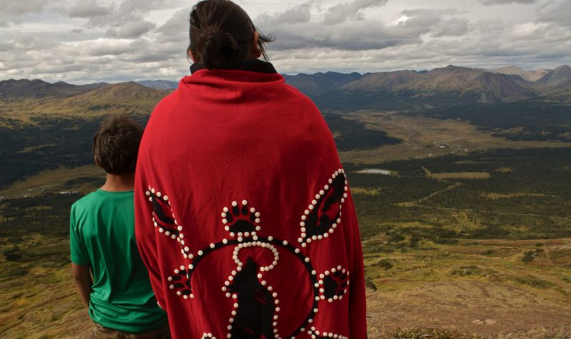 A woman in a red traditional indigenous blanket overlooking a valley with a young boy in a green shirt. both are facing away from the camera.