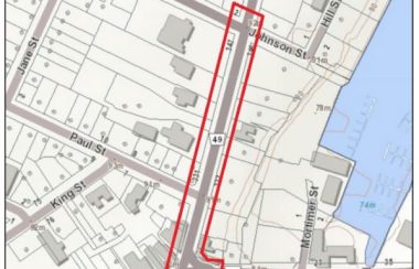 A beige and grey map representing land parcels, buildings, and roads of downtown Picton. A red rectangle-like outline shape highlights the affected area of construction. A blue body of water is off to the right side of the map, representing Picton Bay.