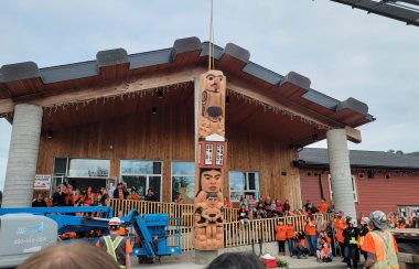 A totem pole being raised in front of a building with surrounding attendees wearing orange shirts
