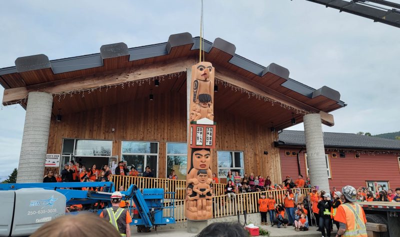 A totem pole being raised in front of a building with surrounding attendees wearing orange shirts