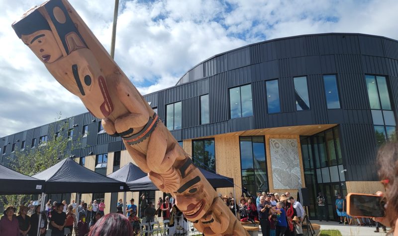 A chain is attached to a wooden totem pole in the process of raising it while people symbolically pull the pole up.