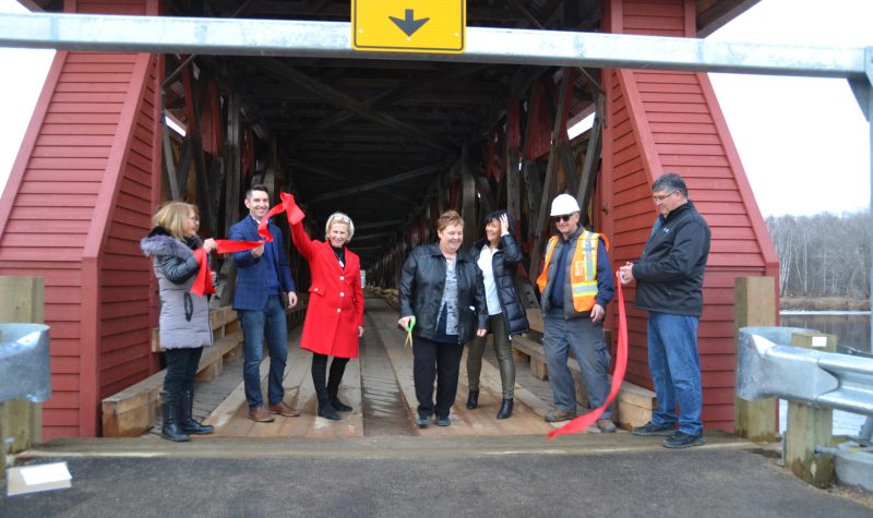 A group of individuals cut a red ribbon in front of a red covered bridge.