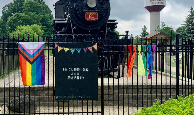 Pride decorations sit on a fence. Trees and a model train sit in the background, with the Palmerston water tower in the distance.