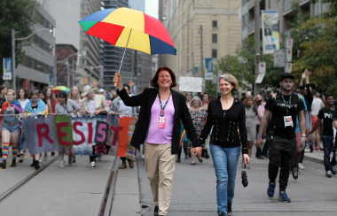 Two people holding a colourful umbrella walk down a street with a crowd holding a banner behind them.