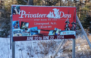 A red road sign for Privateer Days 2014. There is snow on the ground and a sunny woods behind the sign.