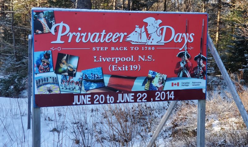 A red road sign for Privateer Days 2014. There is snow on the ground and a sunny woods behind the sign.