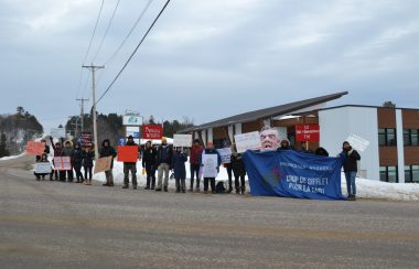 A group of 20 protestors gathers along a highway outside of an office building, holding banners and signs in French denouncing the Quebec government's funding of community organizations.