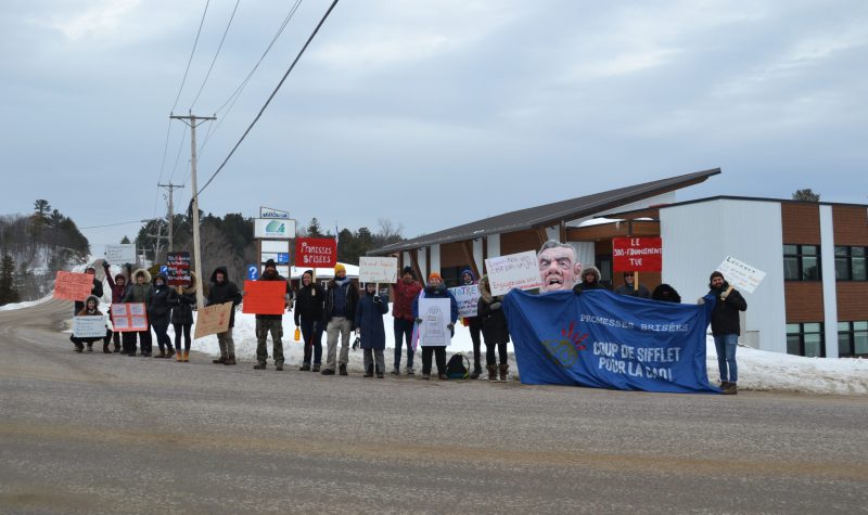 A group of 20 protestors gathers along a highway outside of an office building, holding banners and signs in French denouncing the Quebec government's funding of community organizations.