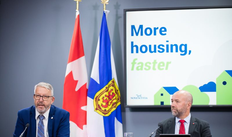 Two men wearing suits sit at a table in front of microphones and flags of Canada and Scotia. There is a corner of a powerpoint projection behind them with the word 'Faster' and three blue and green houses.