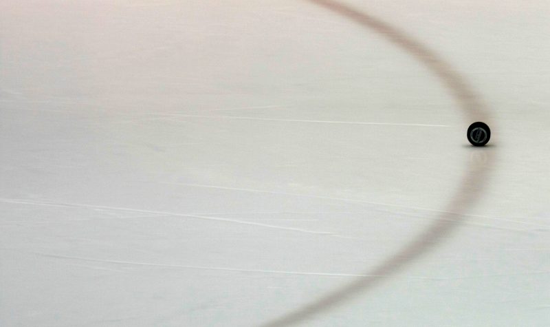 A black hockey puck (with pixelated writing on it) that is standing edgewise on a prepared icy surface. The puck is standing on a red painted line which goes on to depict a half circle, filling the image. As well, a faint reflection of a rink advertisement can be seen along the top border of the image.