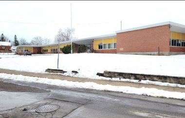 A photo of a school building. In the foreground, a dark and cracked paved parking lot borders a small yard of white snow. Beyond it, in the background is a long square red bricked building with yellow siding surrounding its square windows.