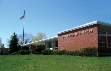 A red brick rectangular building with a Canadian flag flying full beside it. Before it is a field of green grass. A clear blue sky opens up above.