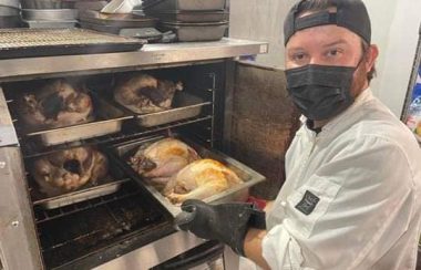 A man wearing a while coat, black baseball cap and white shirt holds a tray of two turkeys in front of an open oven door with three more trays of turkeys inside.