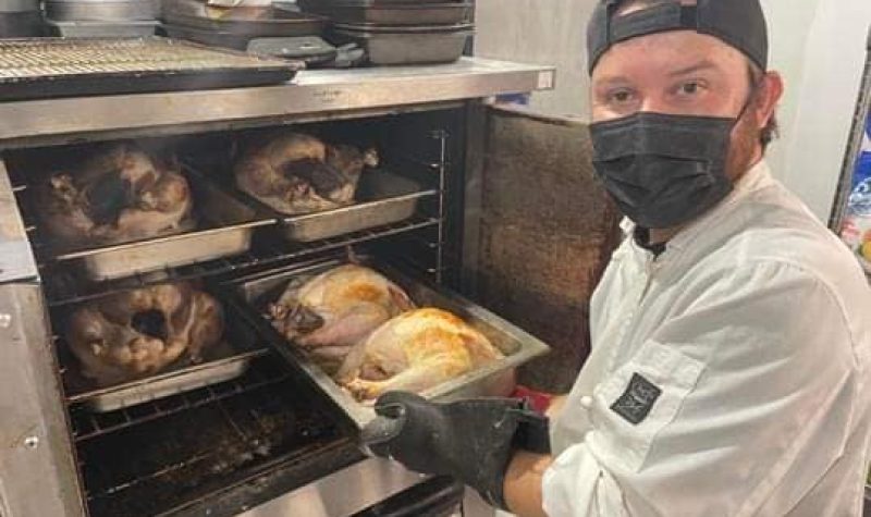 A man wearing a while coat, black baseball cap and white shirt holds a tray of two turkeys in front of an open oven door with three more trays of turkeys inside.