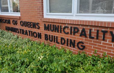 Lettering under two windows and above garden shrubs on a red brick wall which read Region of Queens Municipality Administration Building.