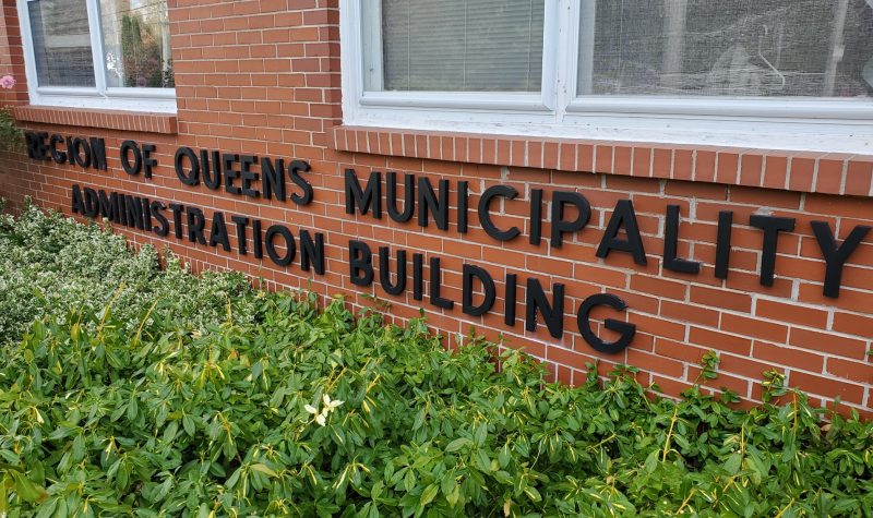 Lettering under two windows and above garden shrubs on a red brick wall which reads 'Region of Queens Municipality Administration Building'.
