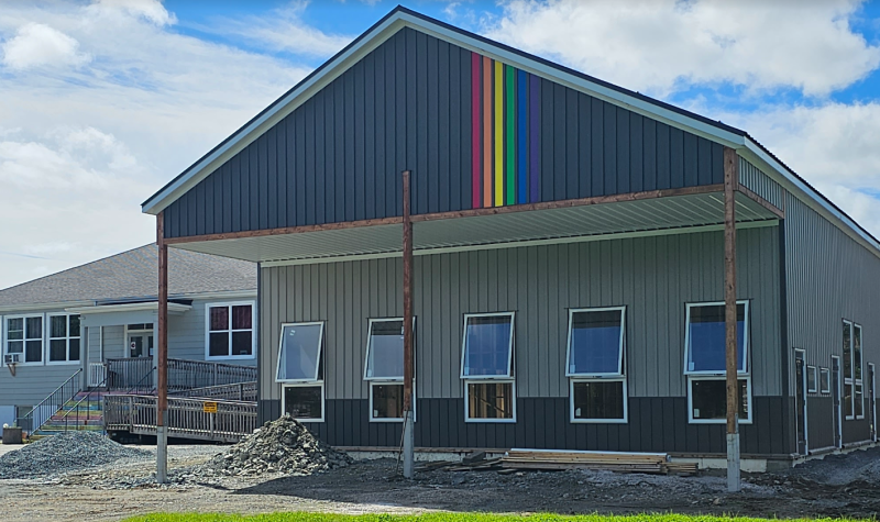 A one story beige and grey building with windows across the front and a rainbow flag near the roofline sits on a parking lot with piles of crushed stone.