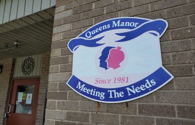 A blue, white and pink exterior sign for Queens Manor. It has two outlines of heads on it, one male and one female, in pink and blue.